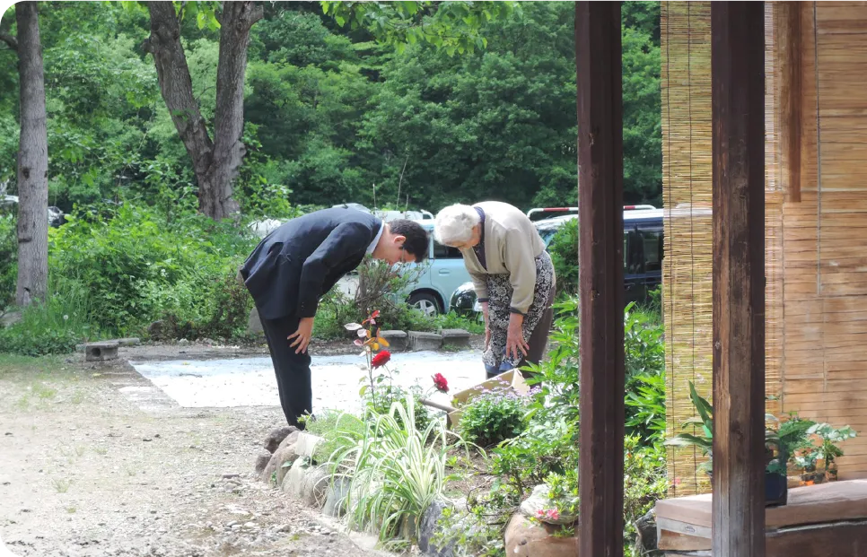 藤原たかし氏が庭でお辞儀をして高齢女性に挨拶している場面